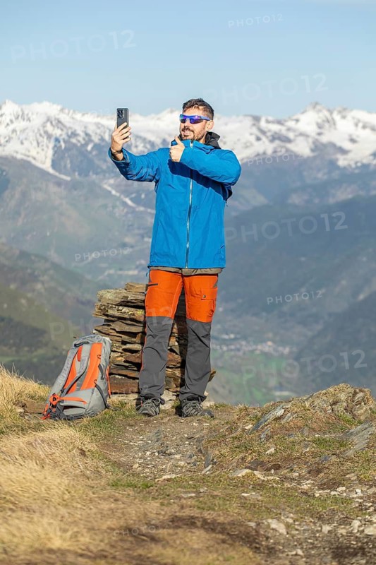 A man, wearing a blue jacket and orange pants, takes a selfie while trekking on a picturesque trail in the Pyrenees. Snow capped mountains beautifully frame the background