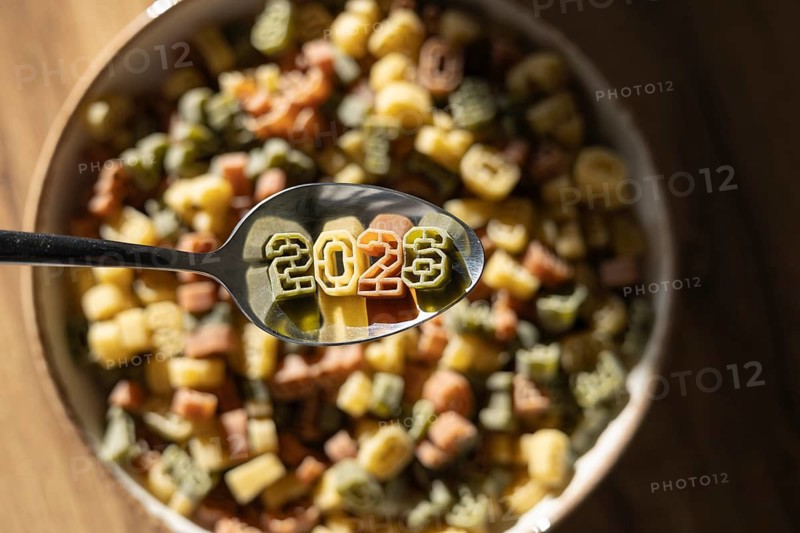 A spoon displaying 2026 in colorful number shaped pasta celebrates the upcoming new year, showcased amidst a bowl of assorted pasta