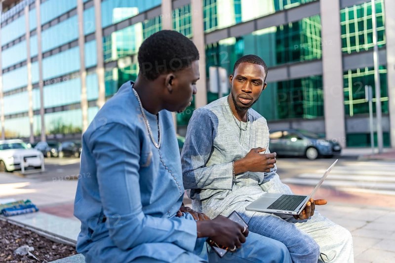 Two senegalese businessmen wearing traditional dashiki attire are discussing business outdoors, using a laptop