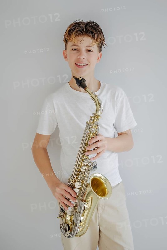 A young musician in a white t shirt smiles while holding a saxophone against a neutral background. The image captures the joy of playing music