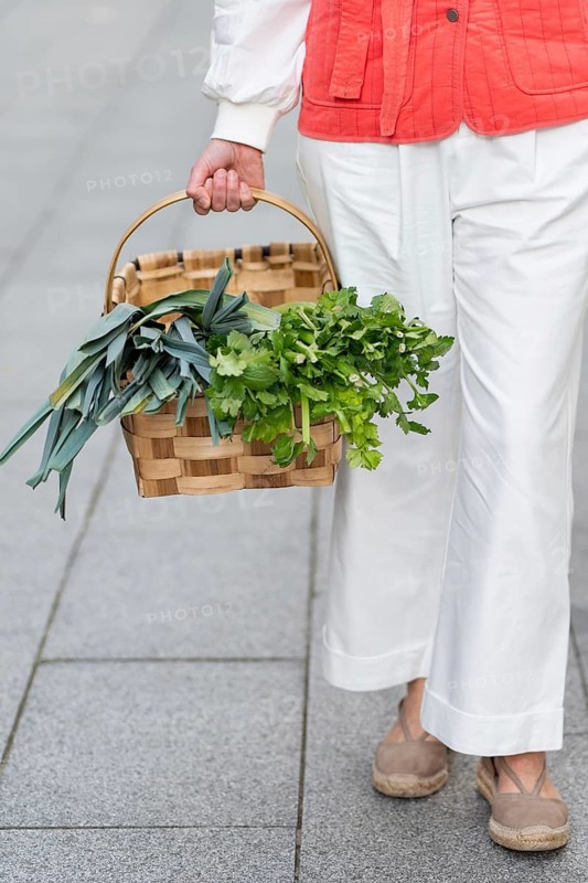 Fashion woman in white pants and red jacket holding a wicker basket filled with fresh herbs, walking on a stone pavement. The scene captures a cozy, outdoor setting