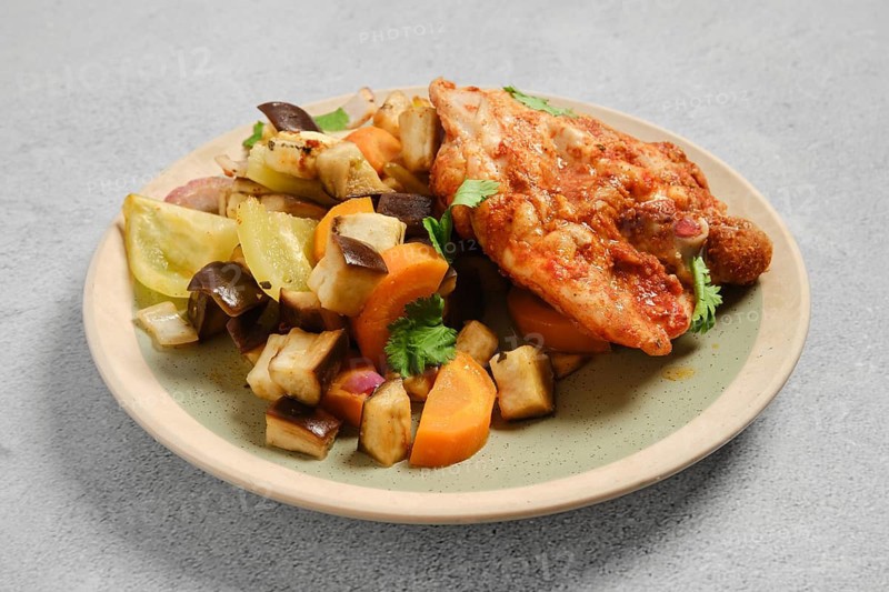 Grilled chicken piece sits on plate next to mix of roasted vegetables including carrots, eggplant and bell peppers. Dish includes fresh herbs for garnish. Setting is a simple light surface