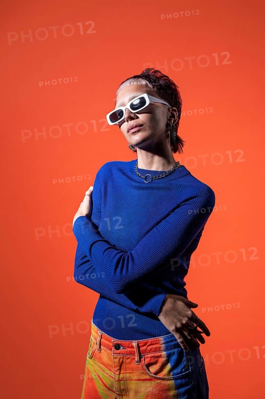 Fashionable young woman wearing vibrant orange and blue trousers, a blue sweater, and chic white sunglasses. Set against a bold orange background, she exudes modern style and confidence