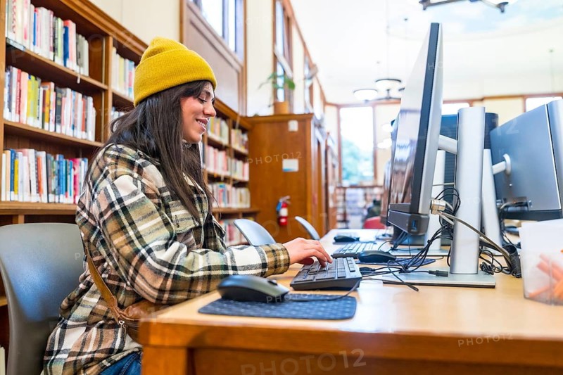 Young millennial woman wearing a beanie and plaid shirt sitting at a computer workstation, actively typing and researching in a traditional public library setting with bookshelves