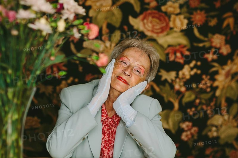 An elegant elderly woman poses with grace in a studio adorned with floral patterns. Her gentle smile and thoughtful expression embody warmth and wisdom