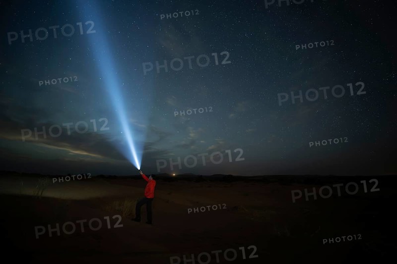 A man with flashlight in a star filled night sky over a Merzouga desert landscape. The faint glow on the horizon create a mystical atmosphere