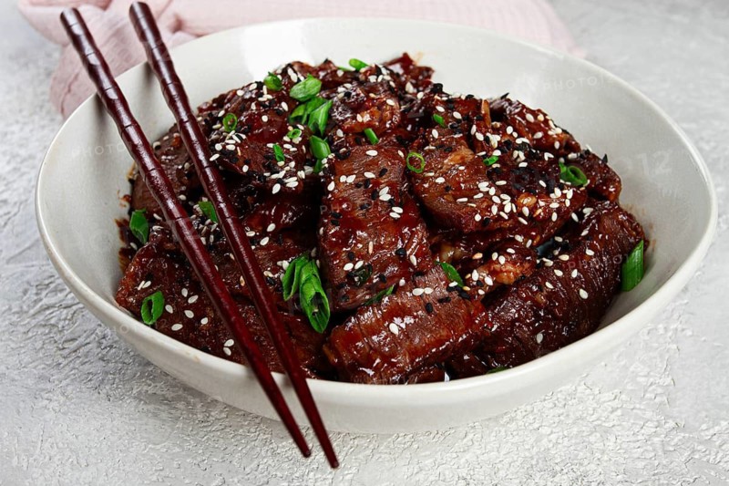Crispy beef, with green onions and sesame seeds, in a bowl with chopsticks, on a textured surface, on a light background, no people