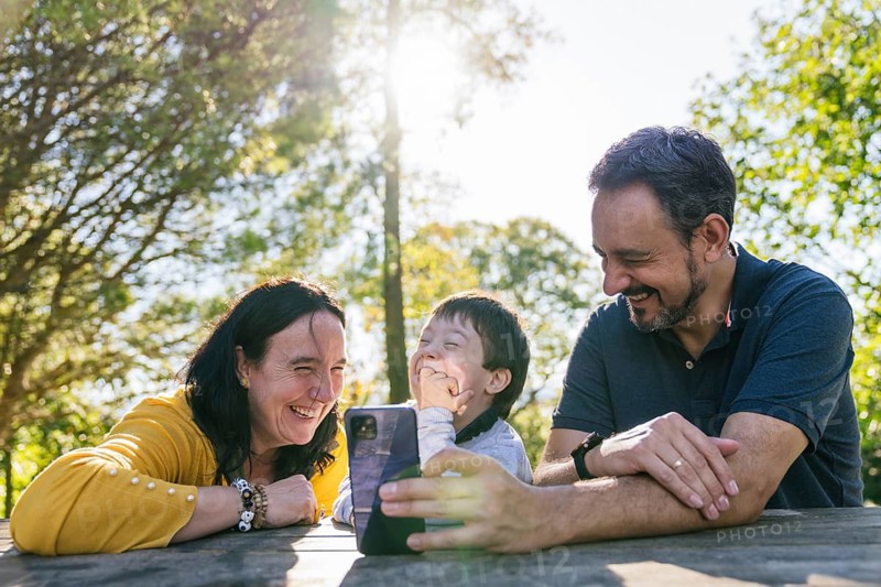 A joyful family takes a selfie at a picnic table with sunlight streaming through the trees. They are outdoors, enjoying quality time and laughter in a serene setting