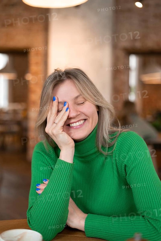 A woman in a green sweater smiles warmly while enjoying breakfast at a cozy cafe. The ambient lighting and background create a welcoming, cheerful atmosphere