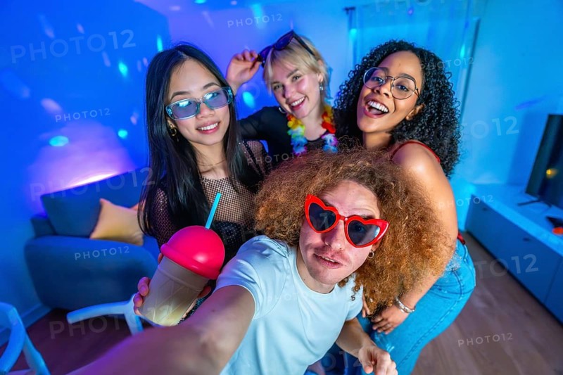 Cheerful multi ethnic friends enjoying a vibrant home disco party, posing for a selfie with colorful lights and refreshing drinks