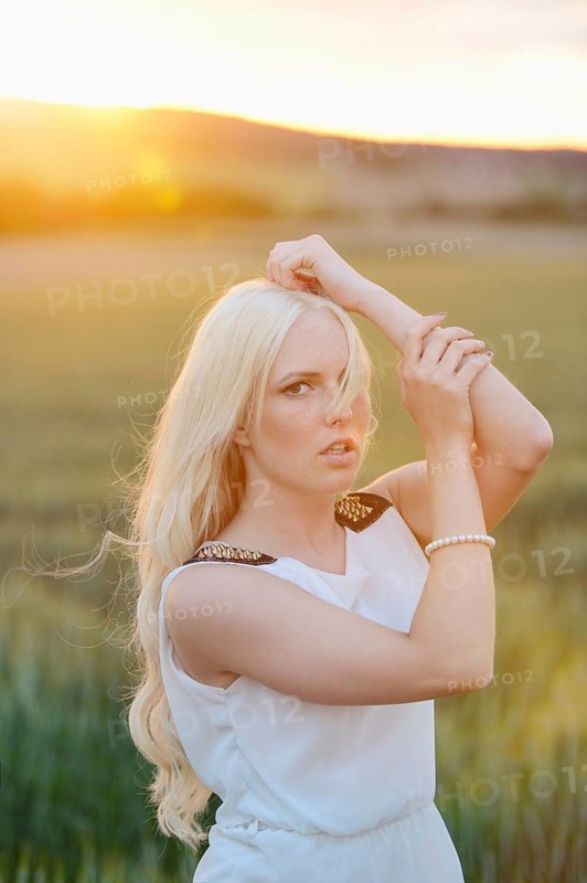 A blonde woman in a white dress poses in a grassy field with a sunset backdrop