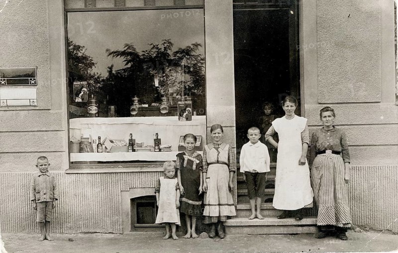 Colonial goods, groceries, children saleswoman, shop, profession, twenties, barefoot, poverty, family standing in front of a shop window displaying goods, window with reflecting trees, around 1914