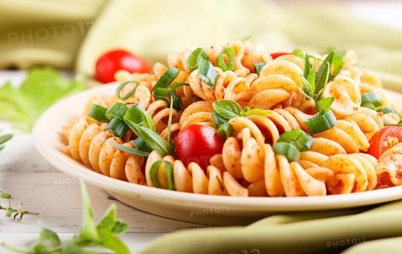 Fusilli pasta with tomato sauce, cherry tomatoes, lettuce and herbs on a white wooden background with green textile. side view, close up, selective focus
