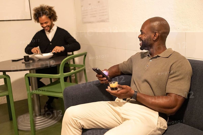 Two men are in a cozy coworking space. One relaxes on a couch with coffee and a smartphone, while another, at a table, enjoys a meal, creating a comfortable work environment