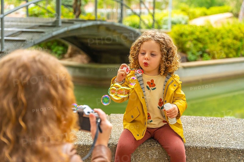 Girl blowing bubbles while mother taking photos of her in a park