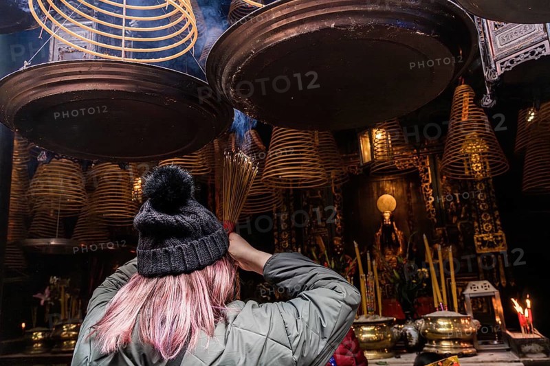 Back view of unrecognizable woman with pink hair lights incense sticks beneath hanging spiral incense at a Taoist and Chinese Buddhist temple in Macau