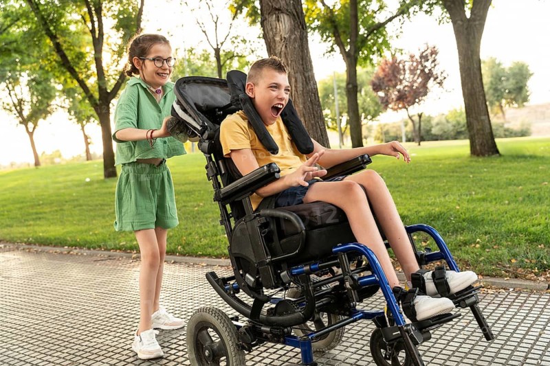 Two children with disabilities enjoy a sunny day in the park. The boy in a wheelchair has cerebral palsy, while the girl with tracheostomy pushes him, both smiling brightly