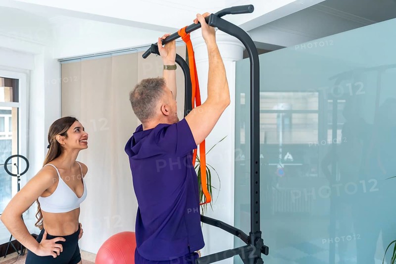 Experienced physiotherapist guiding a mature man performing pull ups with resistance bands on a power tower, focusing on strength building and physical rehabilitation in a clinic setting