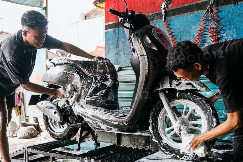 Two asian men meticulously wash a motorcycle, surrounded by soap suds and water. The scene captures the attention to detail and care in motorcycle maintenance