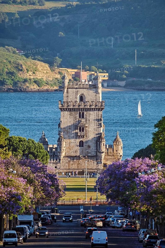 Blooming jacaranda trees line the street as Belem Tower rises above the Tagus River, with sailboats enjoying a tranquil sunset view
