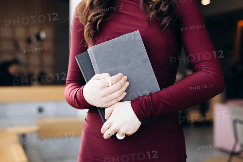 Female student in a maroon dress holding two gray books, standing in a contemporary study room with soft lighting, suggesting a focused and studious atmosphere