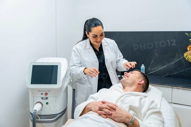 Female cosmetologist in a white lab coat preparing a cheerful male client in a bathrobe for an aesthetic procedure in a clean, modern clinic with specialized equipment