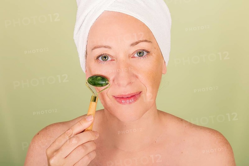 A woman with a towel wrapped around her head uses a jade roller on her face, symbolizing relaxation and self-care against a calming green background