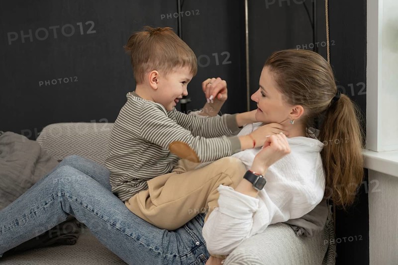 A joyful mother and son share a playful moment on the sofa. The child sits on his mom is lap, both smiling and enjoying quality time in a cozy living room setting