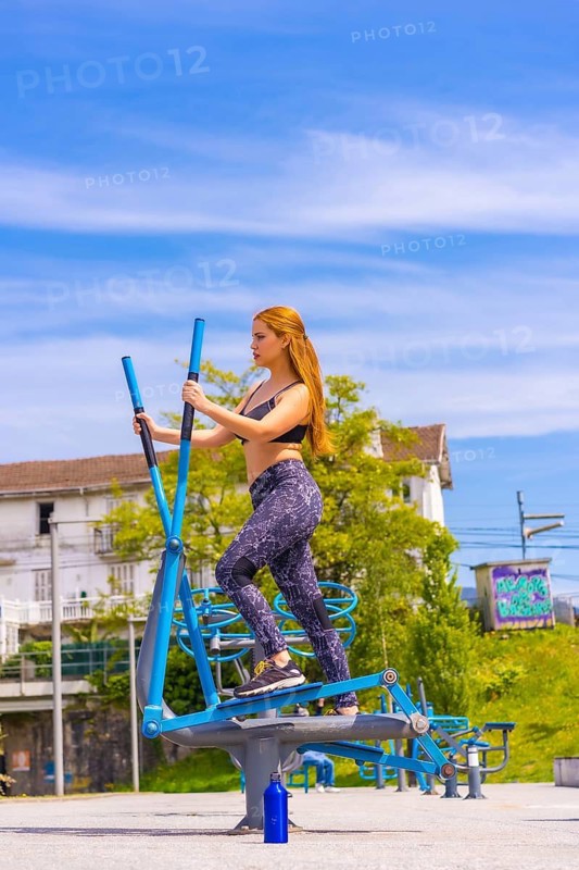 Red-haired woman with purple leggings and black overalls exercising on the machines, sport in the city