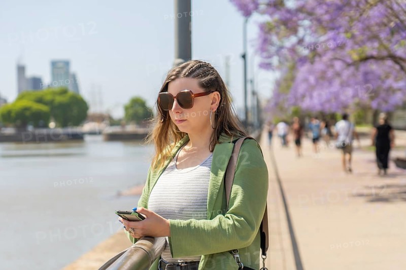 A waist-up shot of a woman wearing sunglasses and a green jacket, focused on typing a message on her smartphone. She is standing near a waterfront surrounded by jacaranda trees and urban scenery