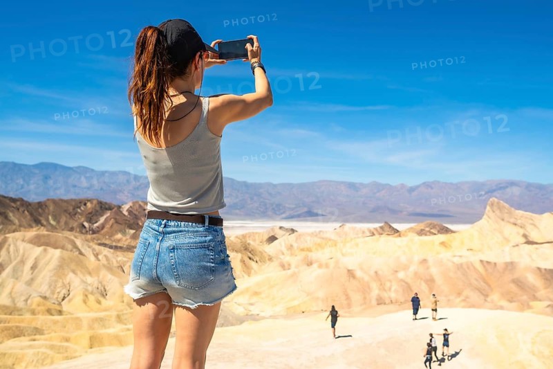 Woman standing at zabriskie point in death valley national park recording the rugged badlands geological formations, distant mountains, and salt flats of badwater basin under a clear blue sky