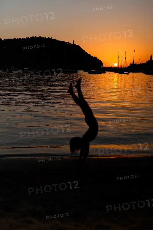 A child silhouette performs a handstand on a sandy beach as the sun sets over a tranquil sea, creating a picturesque and serene scene of evening leisure and play in Soller, Majorca