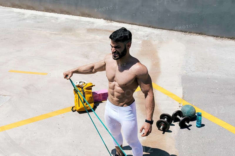 Fit man works out with resistance bands outdoors, showcasing muscular physique in a sunlit urban setting. Exercise equipment like dumbbells and water bottle visible nearby