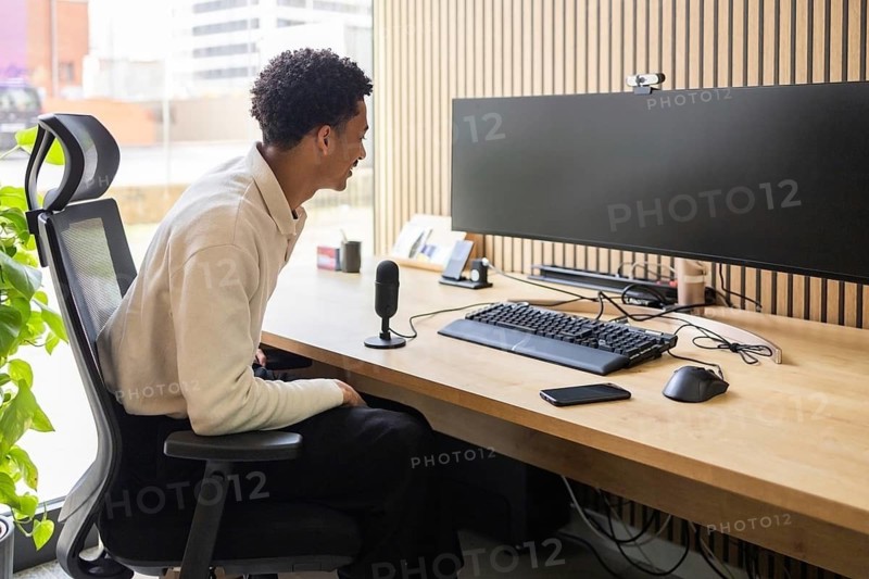 A professional sits comfortably at a modern office desk with a large monitor and microphone. The workspace is organized and well-lit, perfect for productivity and communication