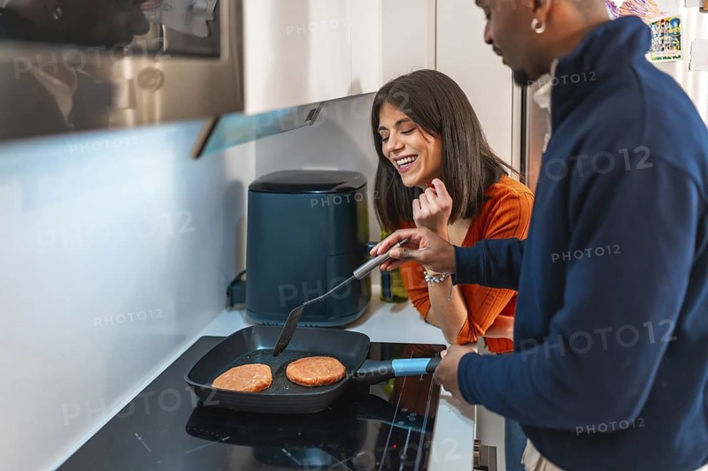 Young multiethnic couple preparing burgers on electric hob in modern kitchen, enjoying their time together