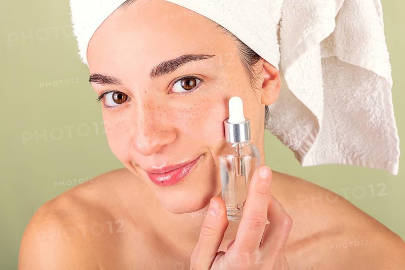 A woman applies a serum to her face, showcasing a skincare and self-care routine. Her hair is wrapped in a towel, emphasizing a fresh, clean approach to beauty