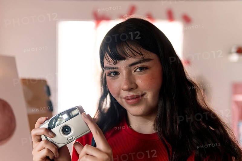 A young woman with long dark hair and a warm smile holds a vintage film camera. She wears a red shirt in a creative studio setting, exuding confidence and artistic expression