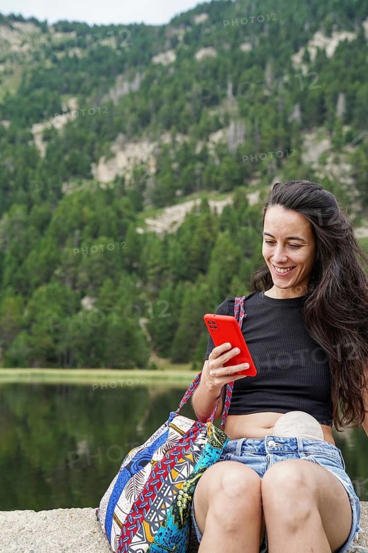 A woman with an ostomy bag is enjoying the outdoors, sitting by a lake, and smiling while looking at her smartphone She radiates happiness and confidence