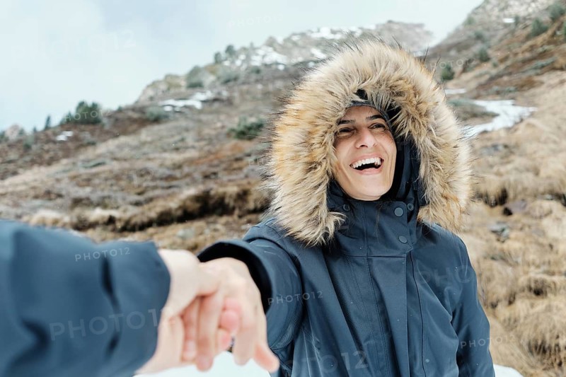 A cheerful woman in a fur-lined hooded coat smiles while holding a hand, set against the snowy slopes of Astun ski resort in the Huesca Pyrenees