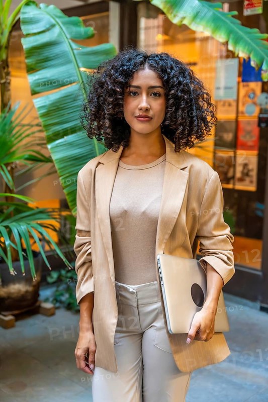 Vertical photo of a casual beauty businesswoman with curly hair standing holding a laptop outside a coworking space