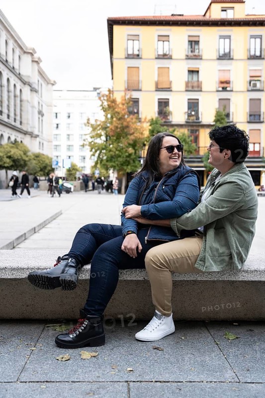 A joyful lesbian couple sitting together in a city square, sharing a moment of love and laughter on a sunny day. Surrounded by people and urban architecture