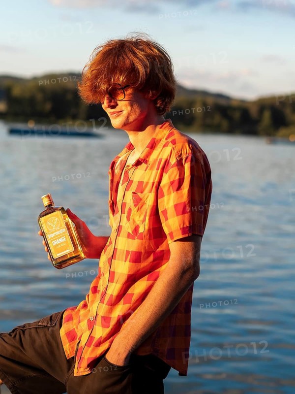 Young man standing by the lake, holding a bottle of alcohol in his hands, herbal liqueur, red hair, orange shirt