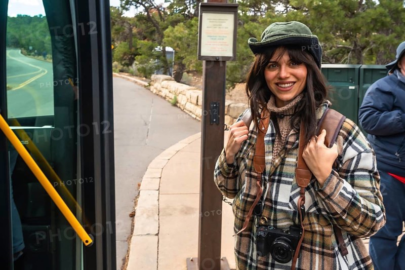 Young woman tourist with backpack and camera smiling while stepping off a public shuttle bus, ready for exploring and sightseeing at a national park stop during her travel journey