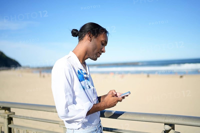 A man stands on a sunny beach, focused on his phone. His relaxed pose and serene surroundings symbolize gay pride and the freedom of the LGBTQIA+ community