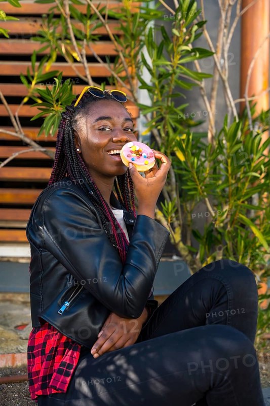 Vertical photo of a cool african teenager eating a doughnut sitting in the street alone