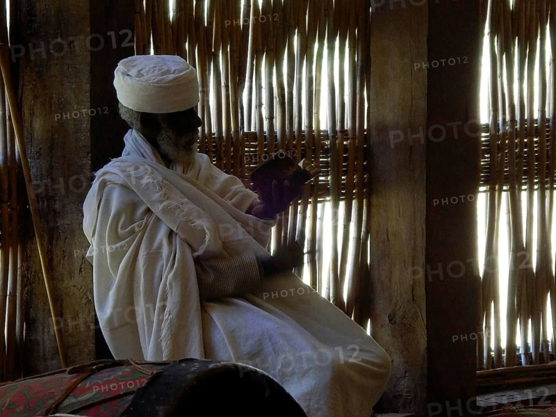 Elderly man wearing traditional garments reading a religious script in a dimly lit room in an Orthodox church, Ethiopia