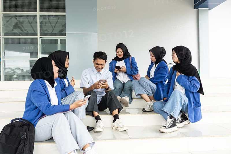 Asian Muslim students in casual attire sit on steps, engaged with smartphones and enjoying each other's company. They share laughter and conversation during a break at school
