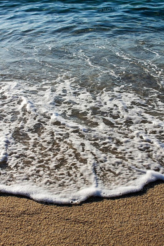 Close-up view of gentle waves washing over a sandy beach, capturing the serene movement of water and foam under the sunlight. Perfect for nature or relaxation themes