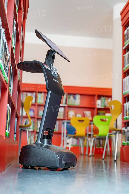 The robot in a library near colourful chairs and shelves, interactive guided tours by robotic personalities from Robolutions, Pforzheim, Germany