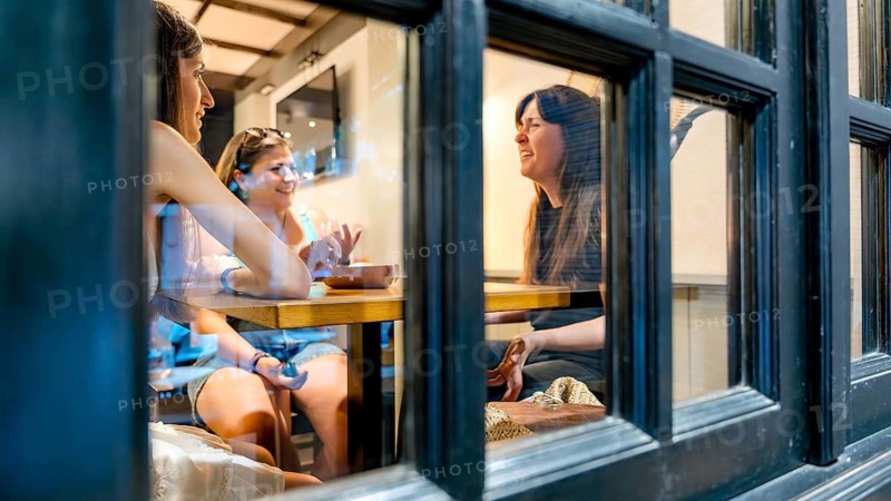 A group of friends sharing laughter and conversation in a cozy restaurant setting. Captured through a window, they enjoy a pleasant dining experience on a sunny day